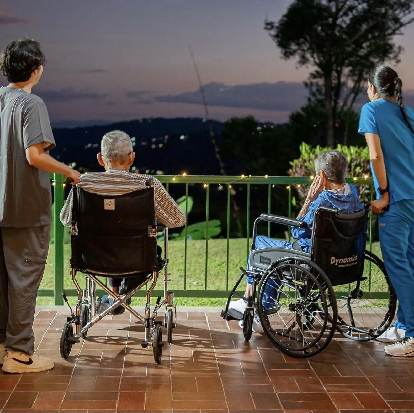 Dos residentes en silla de ruedas miran el atardecer desde la terraza, acompañados por dos cuidadores